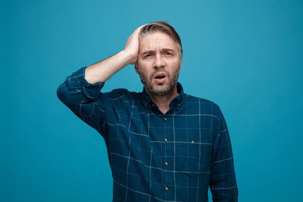 Hombre de mediana edad con cabello gris en camisa de color oscuro que parece confundido sosteniendo la mano en su cabeza por error de pie sobre fondo azul