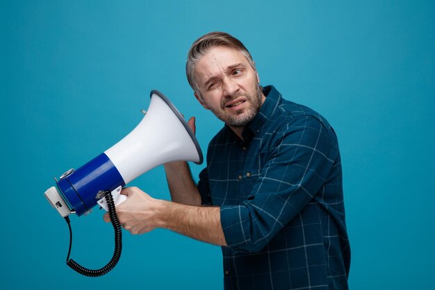 Hombre de mediana edad con cabello gris en camisa de color oscuro sosteniendo megáfono cerca de su oído mirando disgustado de pie sobre fondo azul