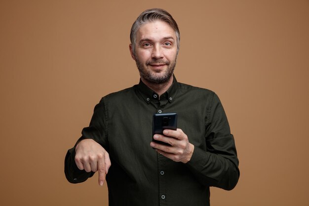 Hombre de mediana edad con cabello gris en camisa de color oscuro sosteniendo un teléfono inteligente apuntando con el dedo índice hacia abajo mirando a la cámara con una sonrisa en la cara de pie sobre fondo marrón