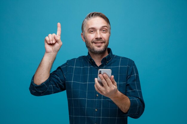 Hombre de mediana edad con cabello gris en camisa de color oscuro sosteniendo un teléfono inteligente apuntando con el dedo índice hacia arriba mirando a la cámara sonriendo feliz y positiva de pie sobre fondo azul