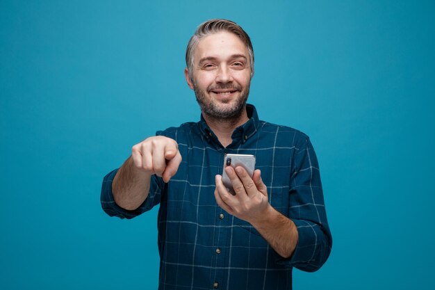 Hombre de mediana edad con cabello gris en camisa de color oscuro sosteniendo un teléfono inteligente apuntando con el dedo índice a la cámara sonriendo confiado feliz y positivo de pie sobre fondo azul