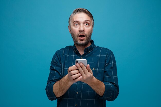 Hombre de mediana edad con cabello gris en camisa de color oscuro sosteniendo un teléfono inteligente mirando a la cámara asombrado y sorprendido de pie sobre fondo azul
