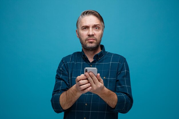 hombre de mediana edad con cabello gris en camisa de color oscuro sosteniendo un teléfono inteligente mirando a la cámara insatisfecho haciendo que la boca irónica frunciera el ceño de pie sobre fondo azul