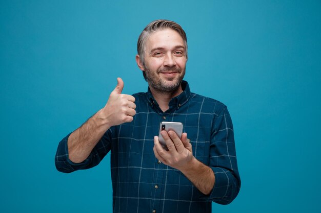 Hombre de mediana edad con cabello gris en camisa de color oscuro sosteniendo un teléfono inteligente mostrando el pulgar hacia arriba mirando a la cámara feliz y alegre sonriendo de pie sobre fondo azul