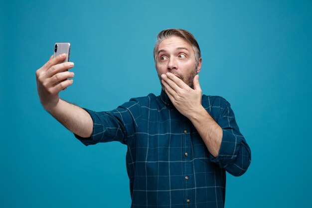 Hombre de mediana edad con cabello gris en camisa de color oscuro sosteniendo un teléfono inteligente con videollamada mirando la pantalla asombrado y sorprendido de pie sobre fondo azul