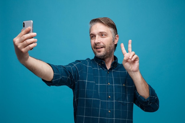 Hombre de mediana edad con cabello gris en camisa de color oscuro con teléfono inteligente con videollamada mirando la pantalla que muestra vsign sonriendo de pie sobre fondo azul