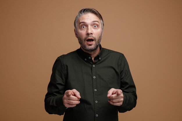 Hombre de mediana edad con cabello gris en camisa verde oscuro mirando a la cámara asombrado y sorprendido señalando con los dedos índices a la cámara de pie sobre fondo marrón