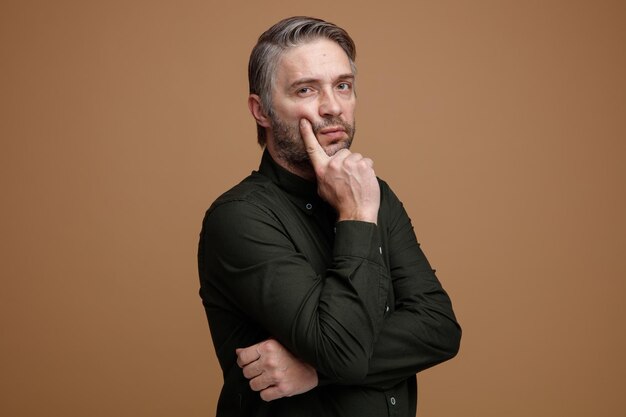 Hombre de mediana edad con cabello gris en camisa verde oscuro mirando a la cámara con expresión pensativa pensando sosteniendo la mano en su barbilla de pie sobre fondo marrón