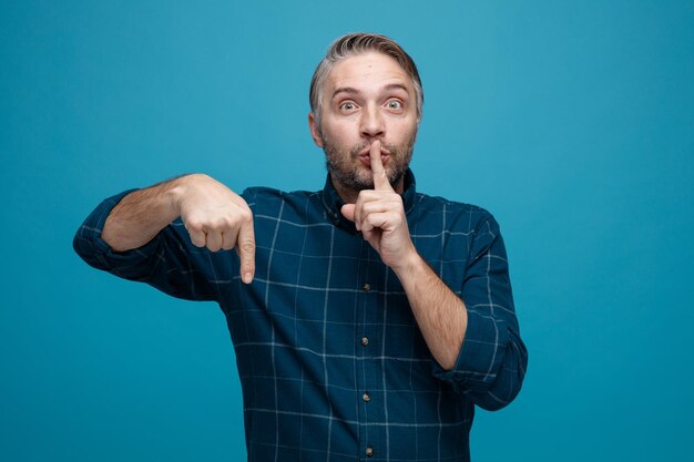 Hombre de mediana edad con cabello gris en pantalones de color oscuro mirando a la cámara con cara seria haciendo gesto de silencio con el dedo en los labios apuntando con el dedo índice hacia abajo de pie sobre fondo azul