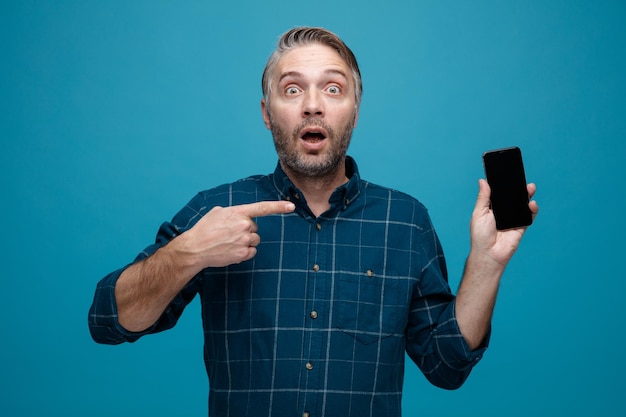 hombre de mediana edad con cabello gris en pantalones de color oscuro que muestra el teléfono inteligente apuntando con el dedo índice mirando a la cámara asombrado y sorprendido de pie sobre fondo azul