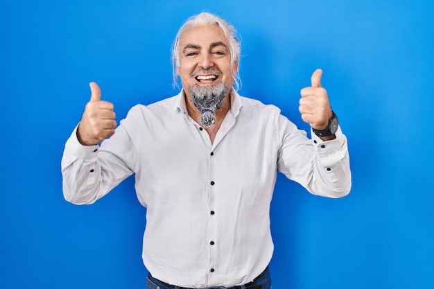 Foto gratuita hombre de mediana edad con cabello gris sobre fondo azul signo de éxito haciendo gesto positivo con los pulgares hacia arriba sonriendo y feliz expresión alegre y gesto de ganador