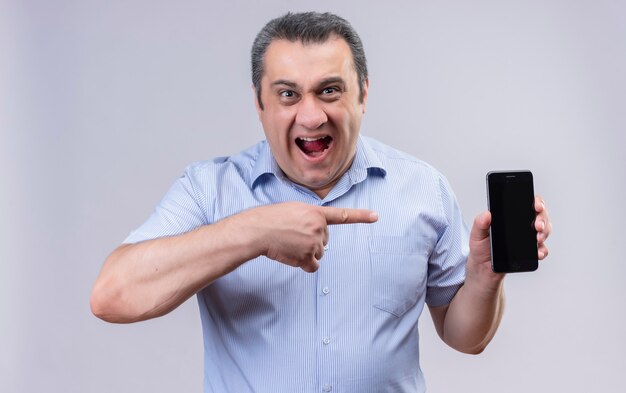 Hombre de mediana edad en camisa azul a rayas verticales manteniendo la boca abierta y señalando con el dedo índice su teléfono móvil mientras está de pie sobre un fondo blanco.