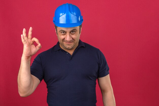 Hombre de mediana edad con camisa polo y casco de seguridad haciendo un signo bien con una sonrisa en la cara sobre la pared rosa aislado