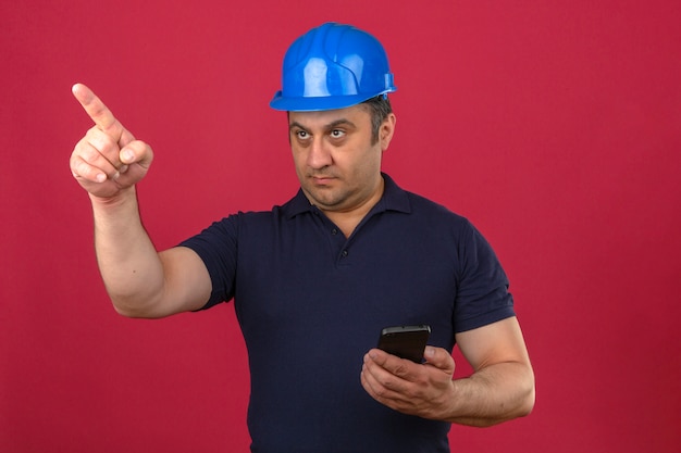 Hombre de mediana edad con camisa polo y casco de seguridad de pie con teléfono inteligente y apuntando con el dedo hacia un lado con cara seria sobre pared rosa aislado