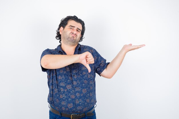 Hombre de mediana edad en camisa sosteniendo algo mientras muestra el pulgar hacia abajo y mirando disgustado, vista frontal.