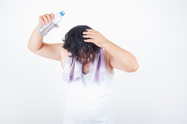 Hombre de mediana edad en camiseta sin mangas, toalla vertiendo agua sobre la cabeza con botella y mirando divertido, vista frontal.