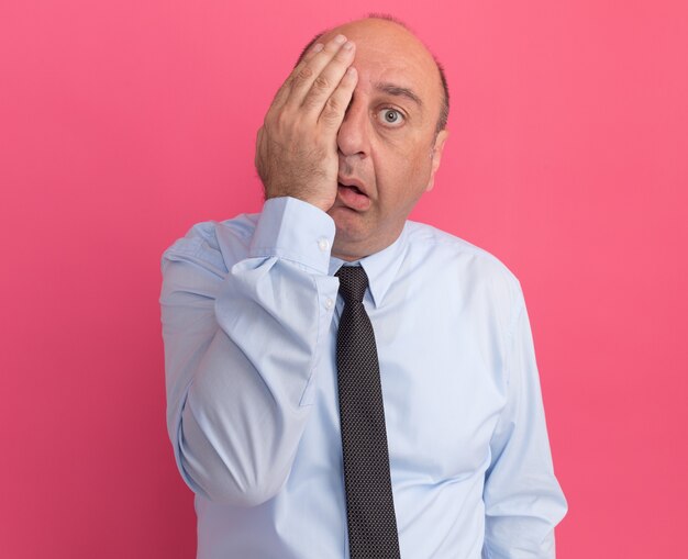 Hombre de mediana edad cansado vestido con camiseta blanca con corbata cubierta de ojos con la mano aislada en la pared rosa