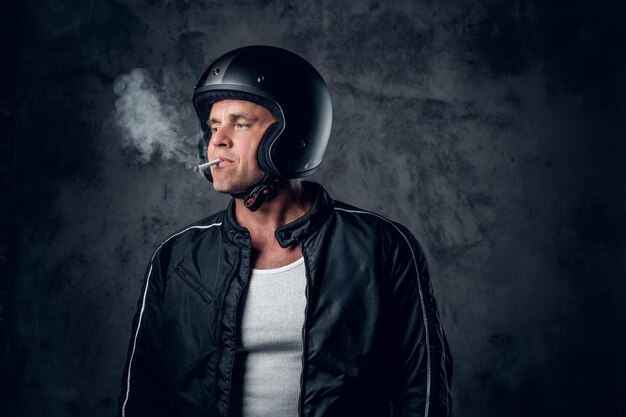 Hombre de mediana edad con casco de motocicleta y chaqueta de cuero fumando un cigarrillo con fondo gris.