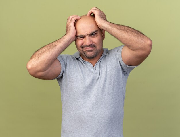 Hombre de mediana edad casual confundido manteniendo las manos en la cabeza aislada en la pared verde oliva