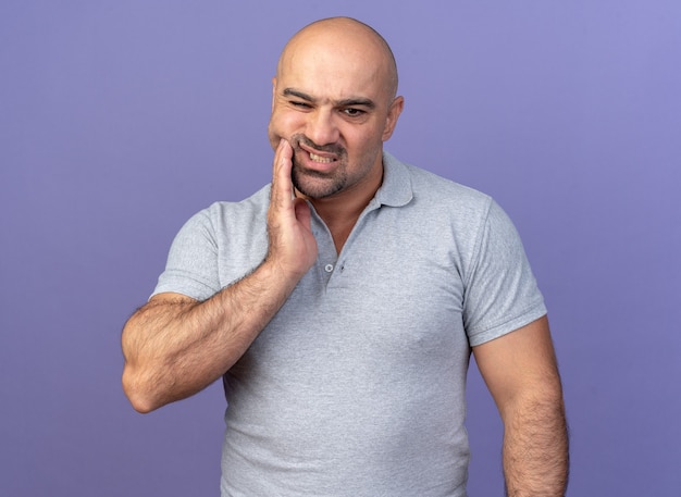 Hombre de mediana edad casual dolorido manteniendo la mano en la mejilla mirando hacia abajo sufriendo de dolor de muelas aislado en la pared púrpura