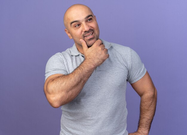 Hombre de mediana edad casual pensativo manteniendo la mano en la barbilla mirando al lado aislado en la pared púrpura