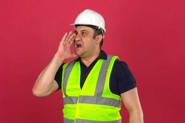 Hombre de mediana edad con chaleco amarillo de construcción y casco de seguridad gritando algo y manteniendo las manos cerca de su boca abierta sobre una pared rosa aislada