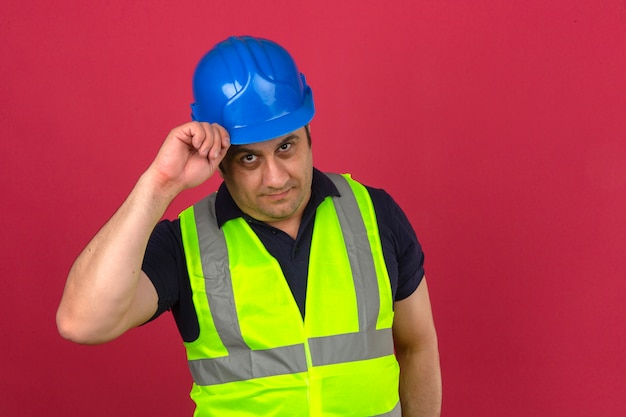 Hombre de mediana edad con chaleco amarillo de construcción y casco de seguridad haciendo gesto de saludo tocando su casco con una sonrisa en la cara sobre la pared rosada aislada
