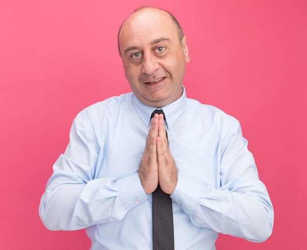 Hombre de mediana edad complacido con camiseta blanca con corbata mostrando gesto de oración aislado en la pared rosa