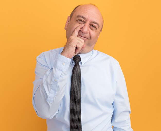Hombre de mediana edad complacido con camiseta blanca con corbata poniendo el dedo en la nariz aislado en la pared naranja