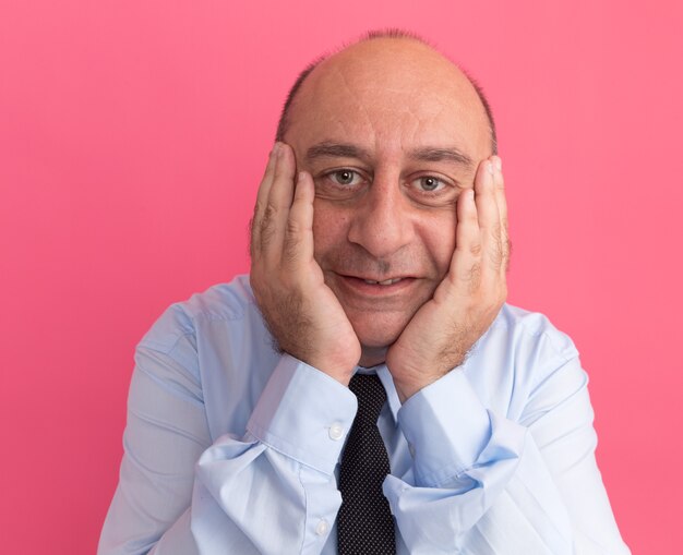 Hombre de mediana edad complacido con camiseta blanca con corbata poniendo las manos en las mejillas aisladas en la pared rosa