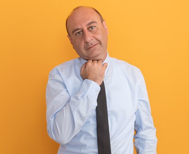 Hombre de mediana edad complacido con camiseta blanca con corbata sujetando el cuello aislado en la pared naranja