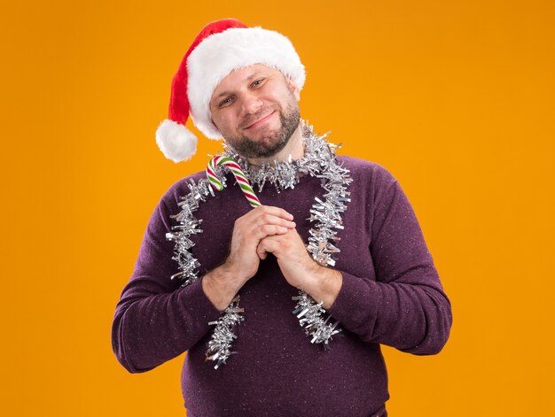 Hombre de mediana edad complacido con gorro de Papá Noel y guirnalda de oropel alrededor del cuello sosteniendo bastón dulce de Navidad mirando a cámara aislada sobre fondo naranja