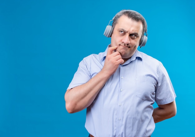 Hombre de mediana edad confundido en camisa a rayas azul con auriculares poniendo la mano en la barbilla para intentar resolver el problema en un espacio azul