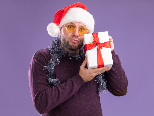 Hombre de mediana edad confundido con gorro de Papá Noel y guirnalda de oropel alrededor del cuello con gafas sosteniendo paquete de regalo aislado en la pared púrpura