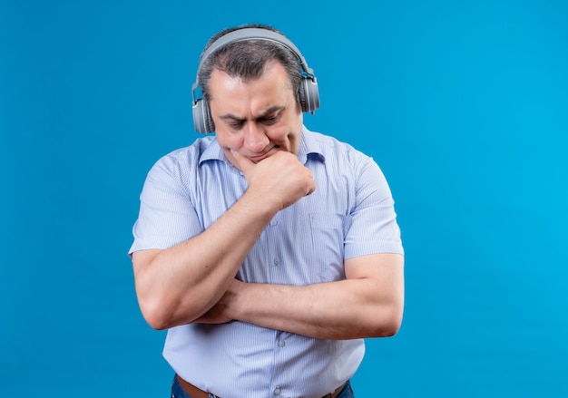 Hombre de mediana edad confundido vestido con camisa a rayas azul puso la mano en la barbilla de pie escuchando algo seriamente con auriculares en un espacio azul