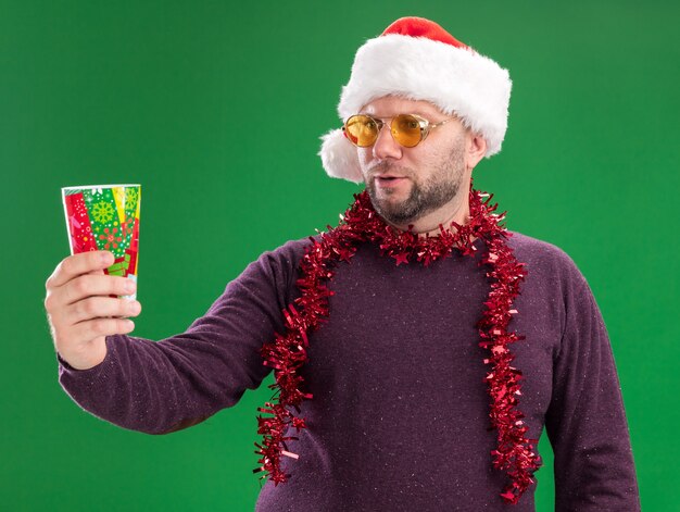 Hombre de mediana edad confundido vestido con gorro de Papá Noel y guirnalda de oropel alrededor del cuello con gafas estirando la taza de Navidad de plástico