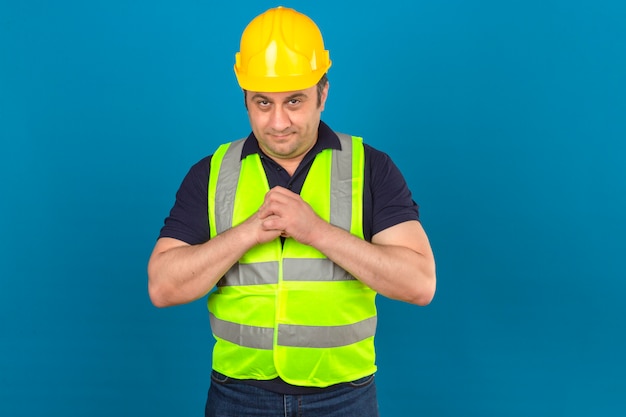 El hombre de mediana edad del constructor que llevaba el chaleco amarillo de la construcción y el casco de seguridad tomados de la mano juntos tramando algo tiene una idea interesante sobre la pared azul aislada