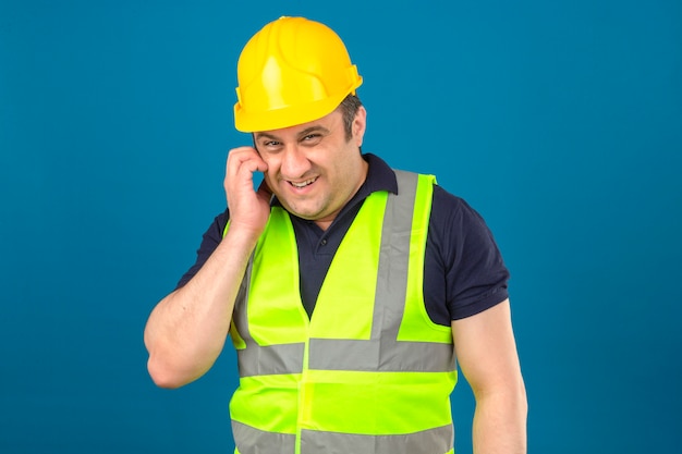 El hombre de mediana edad del constructor que usa el chaleco amarillo de la construcción y el casco de seguridad que rasca la cara que planea algo sonriente astutamente tiene una idea interesante sobre la pared azul aislada