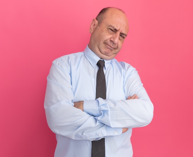Hombre de mediana edad descontento vestido con camiseta blanca con corbata cruzando las manos aisladas en la pared rosa
