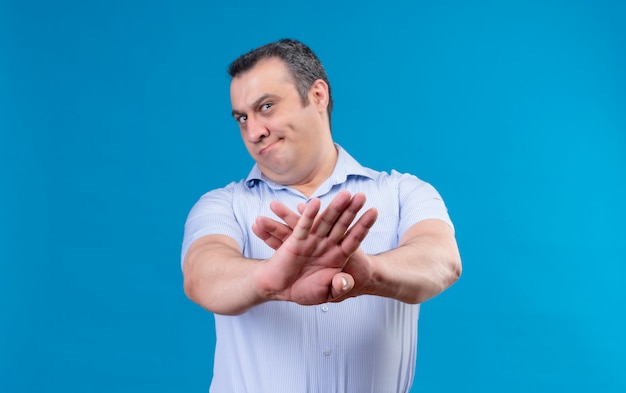 Hombre de mediana edad disgustado en camisa azul de rayas verticales levantando las manos en rechazo en un espacio azul