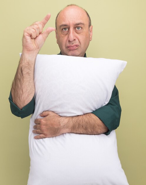 Hombre de mediana edad disgustado vestido con camiseta verde abrazó la almohada mostrando el tamaño aislado en la pared verde oliva