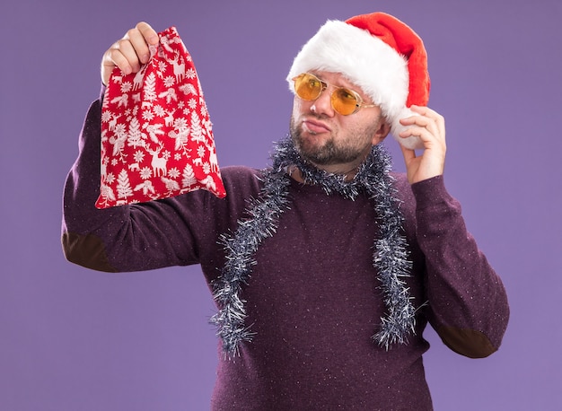 Hombre de mediana edad dudoso con gorro de Papá Noel y guirnalda de oropel alrededor del cuello con gafas sosteniendo y mirando saco de regalo de Navidad agarrando sombrero aislado sobre fondo púrpura