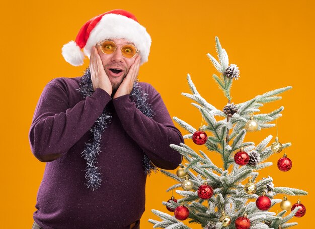 Hombre de mediana edad emocionado con gorro de Papá Noel y guirnalda de oropel alrededor del cuello con gafas de pie cerca del árbol de Navidad decorado manteniendo las manos en la cara mirando a cámara aislada sobre fondo naranja