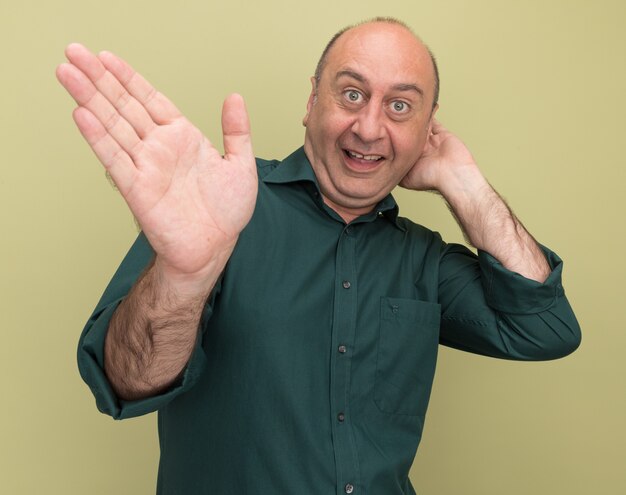 Hombre de mediana edad emocionado con puntos de camiseta verde con la mano al lado poniendo la mano detrás de la cabeza aislada en la pared verde oliva