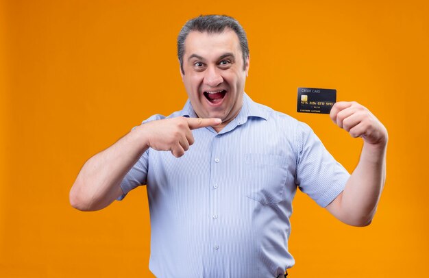 Hombre de mediana edad emocionado vestido con puntos de camisa de rayas verticales azules con tarjeta de crédito de dedo índice manteniendo la boca abierta