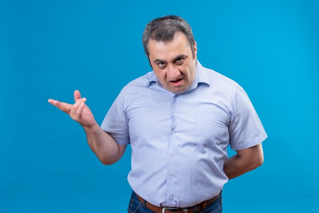 Hombre de mediana edad enojado y serio en camisa a rayas azul apuntando con la mano preguntando qué en un espacio azul