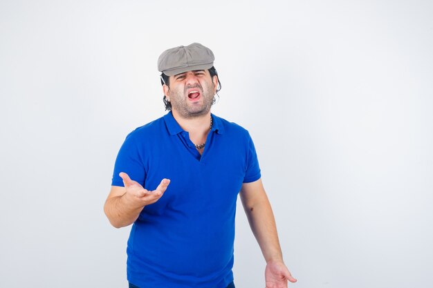 Hombre de mediana edad estirando la mano de manera interrogante en camiseta de polo, sombrero de hiedra y mirando enojado, vista frontal.