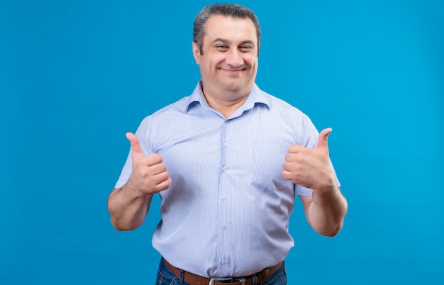 Hombre de mediana edad feliz y emocionado con camisa azul mostrando con ambas manos pulgares hacia arriba como gesto en un espacio azul
