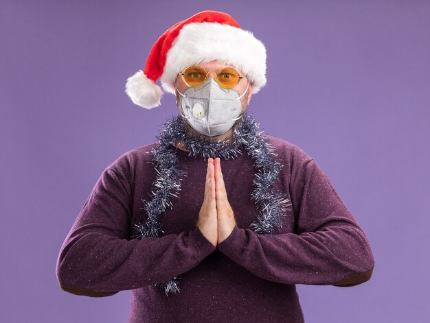 Hombre de mediana edad con gorro de Papá Noel y guirnalda de oropel alrededor del cuello con gafas y máscara protectora manteniendo las manos juntas aisladas en la pared púrpura