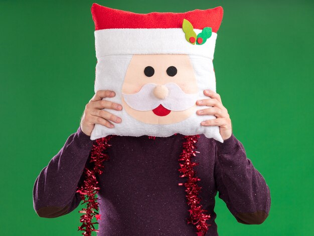 Hombre de mediana edad con gorro de Papá Noel y guirnalda de oropel alrededor del cuello con gafas sosteniendo la almohada de santa claus delante de la cara aislada en la pared verde